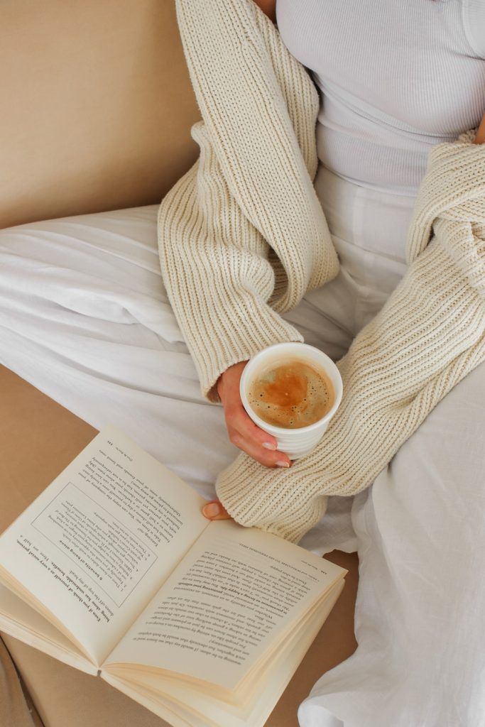 Person wearing a cozy beige sweater holding a cup of coffee next to an open book on a couch.