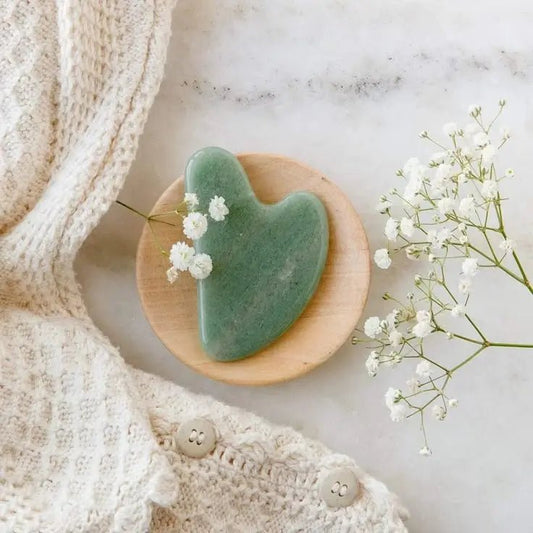 A green jade stone facial massage tool lay flat on flowers and marble background with linen draped on corner of image.